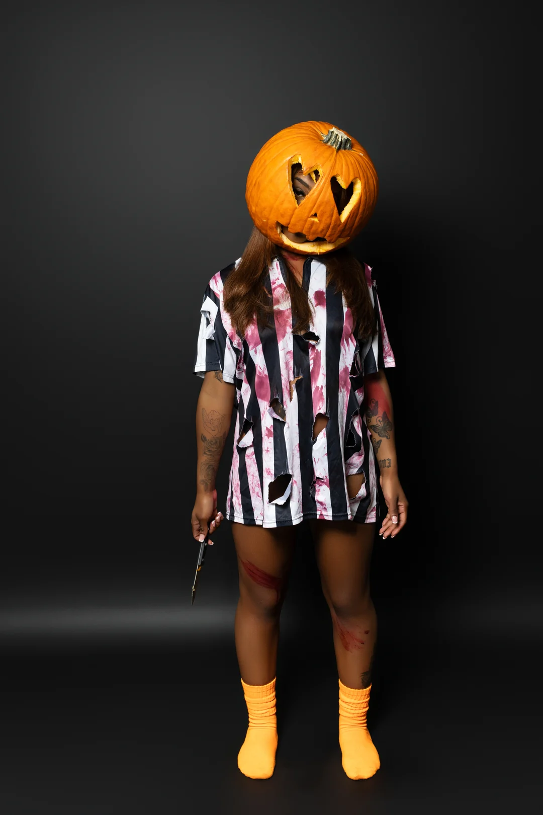 A person wearing a carved pumpkin head, striped shirt and yellow socks poses against a charcoal backdrop.